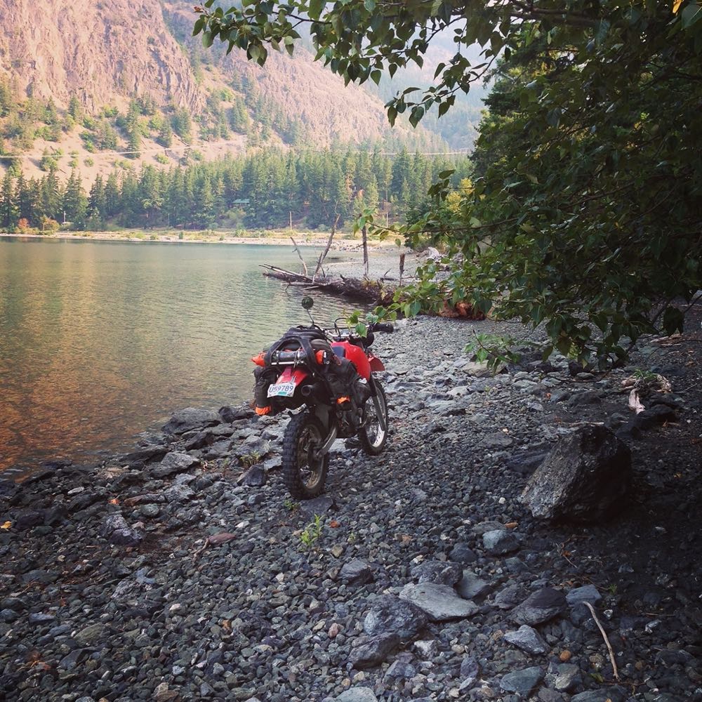 Adventure motorcycle parked on a rocky lakeshore under overhanging trees, still water and forested slopes across the lake