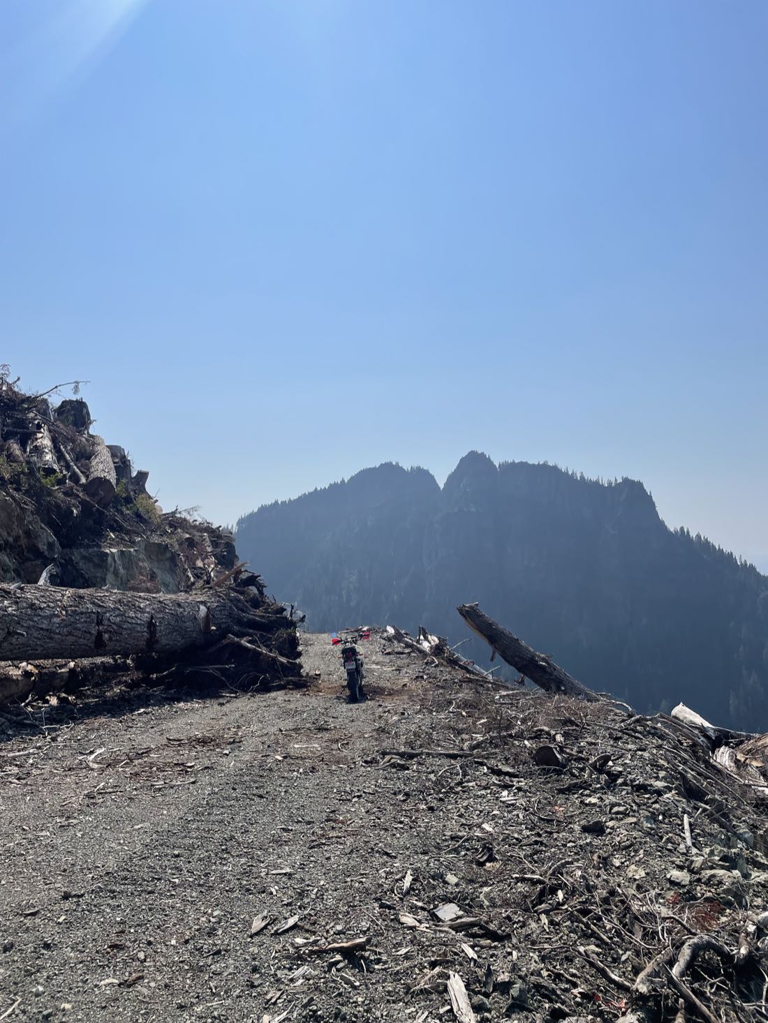 Motorcycle stopped on a high logging road below jagged rock peaks on Vancouver Island, morning haze in the valley