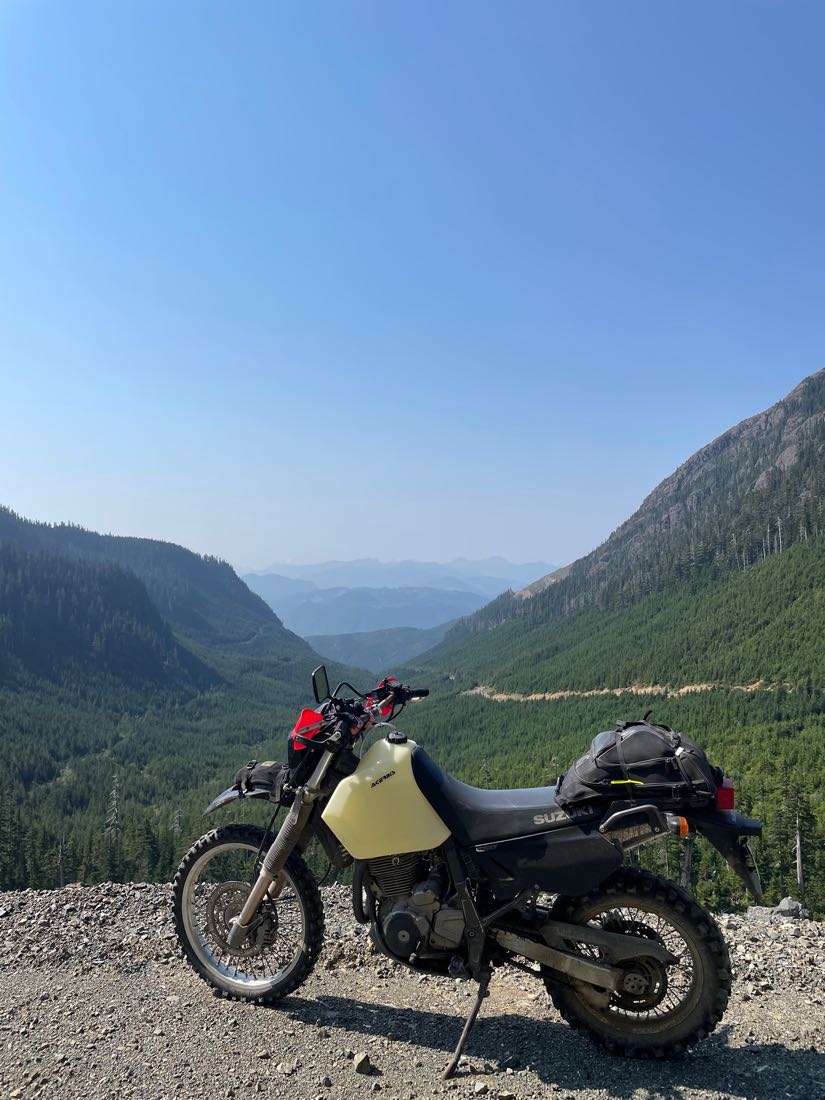 Dual-sport motorcycle parked on a gravel viewpoint high above a forested valley on Vancouver Island, mountain ridges fading into the distance